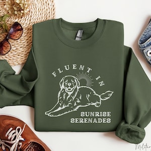 Puede incluir: Una sudadera verde con un gráfico blanco de un perro y el texto "Fluent in Sunrise Serenades".