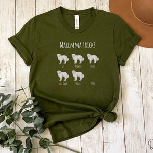 Pode incluir: Camiseta verde oliva com o texto "Maremma Tricks" e ilustra&ccedil;&otilde;es de um c&atilde;o pastor maremmano realizando v&aacute;rios truques, incluindo sentar, deitar, sacudir, rolar, buscar e ficar.