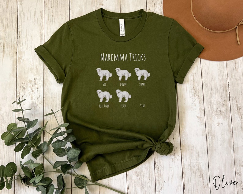 Pode incluir: Camiseta verde oliva com o texto "Maremma Tricks" e ilustra&ccedil;&otilde;es de um c&atilde;o pastor maremmano realizando v&aacute;rios truques, incluindo sentar, deitar, sacudir, rolar, buscar e ficar.