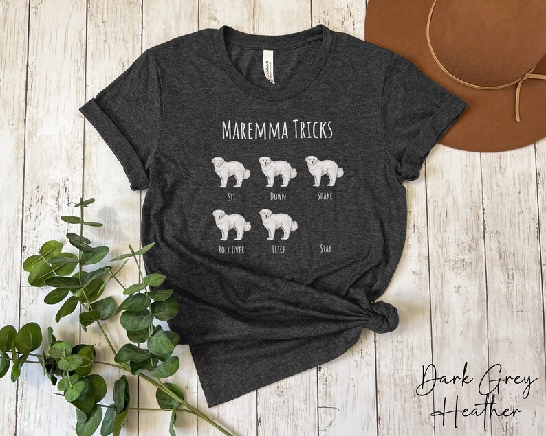 Pode incluir: Camiseta cinza escuro heather com uma ilustra&ccedil;&atilde;o de um c&atilde;o Maremma branco e o texto "Maremma Tricks". O c&atilde;o &eacute; mostrado em diferentes poses, com o truque correspondente abaixo de cada pose: sentar, deitar, sacudir, rolar, buscar, ficar.