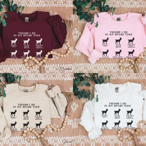May include: Four sweatshirts in maroon, light pink, sand, and white are displayed. Each sweatshirt has the text "THINGS I DO IN MY SPARE TIME" with goat illustrations. The sweatshirts are on a textured surface with decorative beads.