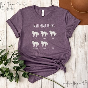Pode incluir: Camiseta roxa heather com um gr&aacute;fico branco de um c&atilde;o pastor maremmano e o texto "MAREMMA TRICKS". O gr&aacute;fico mostra o c&atilde;o realizando diferentes truques, incluindo sentar, deitar, sacudir, rolar, buscar e ficar.