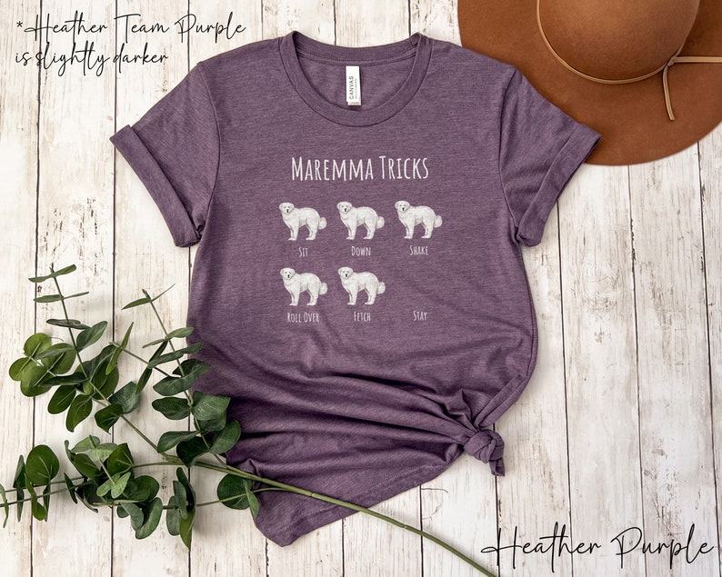 Pode incluir: Camiseta roxa heather com um gr&aacute;fico branco de um c&atilde;o pastor maremmano e o texto "MAREMMA TRICKS". O gr&aacute;fico mostra o c&atilde;o realizando diferentes truques, incluindo sentar, deitar, sacudir, rolar, buscar e ficar.