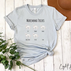 Pode incluir: Camiseta azul claro com o texto "Maremma Tricks" e ilustra&ccedil;&otilde;es de um c&atilde;o branco realizando v&aacute;rios truques, incluindo sentar, deitar, sacudir, rolar, buscar e ficar.