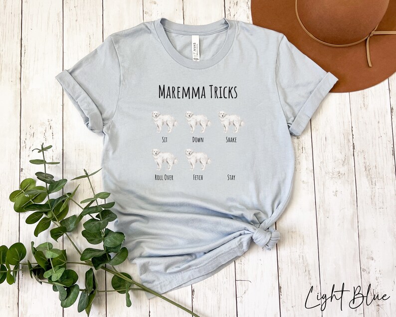 Pode incluir: Camiseta azul claro com o texto "Maremma Tricks" e ilustra&ccedil;&otilde;es de um c&atilde;o branco realizando v&aacute;rios truques, incluindo sentar, deitar, sacudir, rolar, buscar e ficar.