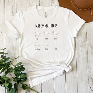 Pode incluir: Camiseta branca com o texto "Maremma Tricks" e ilustra&ccedil;&otilde;es de um c&atilde;o realizando v&aacute;rios truques, incluindo sentar, deitar, dar a pata, rolar, buscar e ficar.