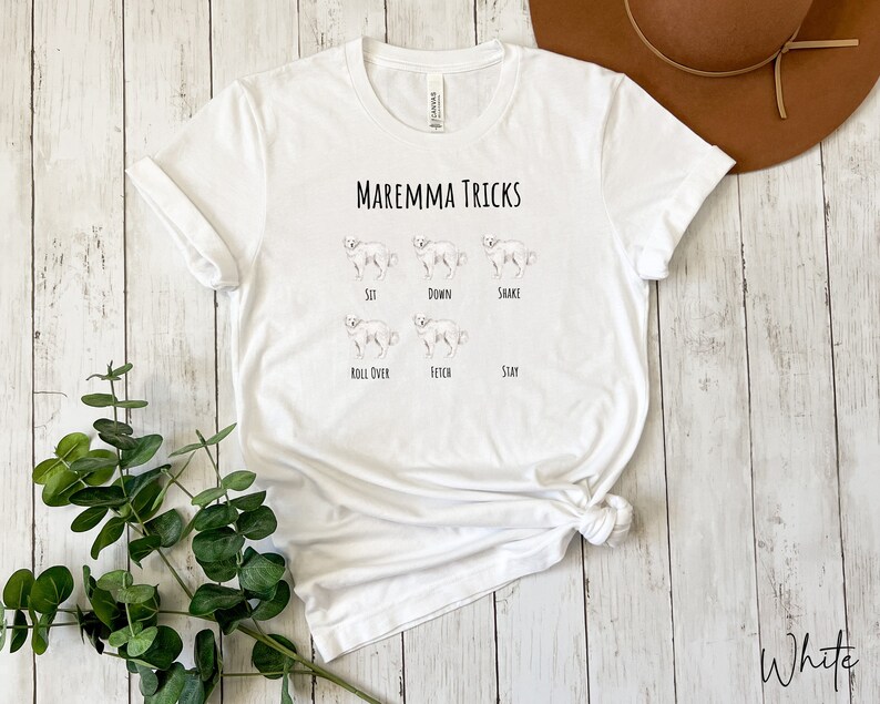Pode incluir: Camiseta branca com o texto "Maremma Tricks" e ilustra&ccedil;&otilde;es de um c&atilde;o realizando v&aacute;rios truques, incluindo sentar, deitar, dar a pata, rolar, buscar e ficar.