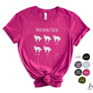 Pode incluir: Uma camiseta rosa com o texto "MAREMMA TRICKS" e ilustra&ccedil;&otilde;es de um c&atilde;o branco realizando v&aacute;rios truques, incluindo sentar, deitar, sacudir, rolar, buscar e ficar.
