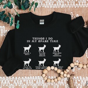 May include: Black long-sleeve sweatshirt with white text and goat illustrations. The text reads "THINGS I DO IN MY SPARE TIME" with phrases like "CHASE A GOAT" and "TALK TO MY GOATS". The word "Black" is written in cursive at the bottom.