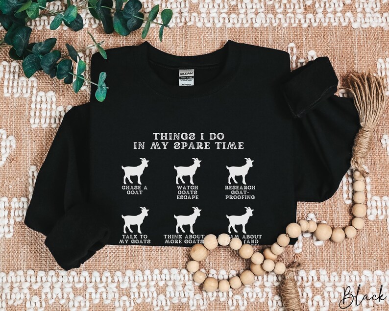 May include: Black long-sleeve sweatshirt with white text and goat illustrations. The text reads "THINGS I DO IN MY SPARE TIME" with phrases like "CHASE A GOAT" and "TALK TO MY GOATS". The word "Black" is written in cursive at the bottom.