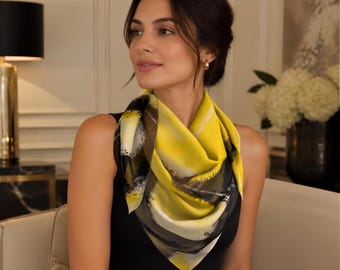 May include: A square silk scarf featuring a vibrant yellow and black abstract brushstroke pattern. The scarf is folded and draped around the neck, showcasing a smooth, lightweight fabric with a glossy finish and dark, sweeping artistic lines.