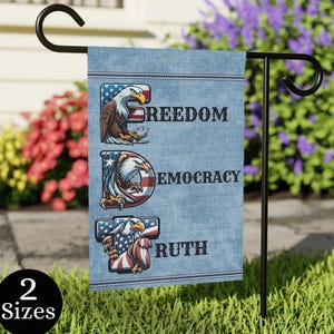 May include: A denim-textured garden flag with the words FREEDOM, DEMOCRACY, and TRUTH, each letter featuring an American bald eagle and the US flag. The flag is displayed on a black metal stand in a garden setting. The flag is available in 2 sizes.