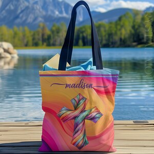 May include: A colorful tote bag with a vibrant, swirling design in shades of orange, pink, and purple. The bag features a rainbow-colored cross and the name "madison" in a cursive font. It has black handles and appears to be on a wooden dock.