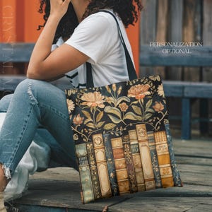May include: Black tote bag featuring a floral and book design. The bag has a dark background with orange and cream-colored flowers and green leaves. Below the flowers are illustrated books with gold lettering. The bag has black straps and the text "PERSONALIZATION OPTIONAL".