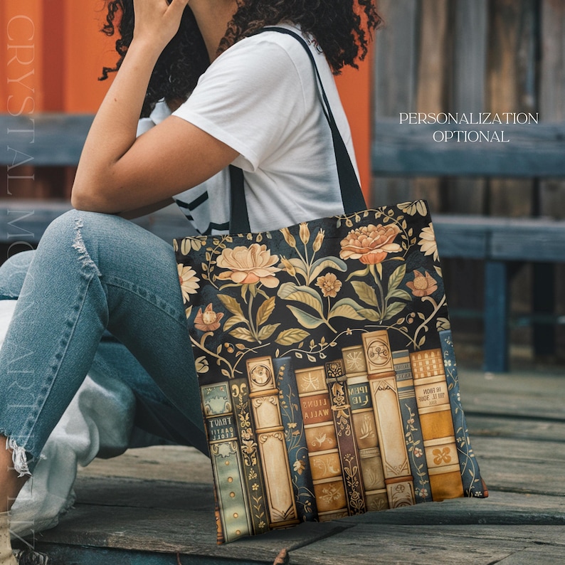 May include: Black tote bag featuring a floral and book design. The bag has a dark background with orange and cream-colored flowers and green leaves. Below the flowers are illustrated books with gold lettering. The bag has black straps and the text "PERSONALIZATION OPTIONAL".