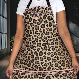 May include: A brown and black leopard print apron with a pink and white trim. The apron has the text "chef nina" in black cursive with a pink heart above the text.