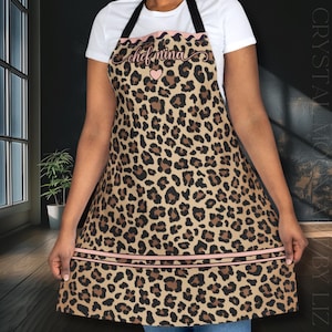 May include: A leopard print apron featuring black straps, a pink trim along the top and bottom, and the text "chef nina" in cursive script above a small pink heart. The apron has a tan base with dark brown and black animal print spots.