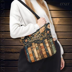 May include: A tote bag with a floral and book design. The bag features the text "Paisley Dark Romance Book Club" and measures 13 inches by 13 inches. The bag has a black strap and a dark background with floral and book illustrations.