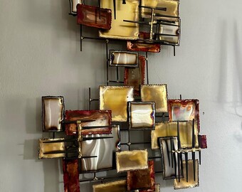 May include: A large abstract wall sculpture made of metal. The sculpture is composed of many rectangular pieces of metal in shades of gold, brown, and red. The pieces are arranged in a random pattern and are held together by thin metal rods.