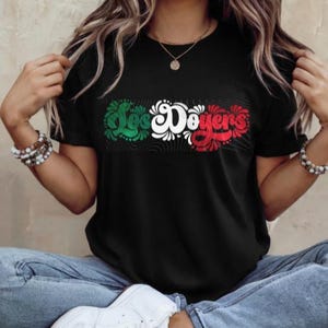 May include: A black t-shirt featuring a graphic of the words "Los Doyers" in green, white and red lettering. The lettering is stylised with swirls and floral designs.