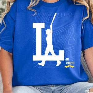 Op de afbeelding: Een koninklijk blauw t-shirt met een wit silhouet van een honkballer die een bal slaat. De speler staat voor een witte "L.A." met een gele "10 2 OUTS" grafiek eronder.