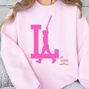 Op de afbeelding: Roze sweatshirt met een roze afbeelding van een honkballer die een bal slaat met de letter "L" op de achtergrond. De afbeelding bevat ook de tekst "10 OUTS".