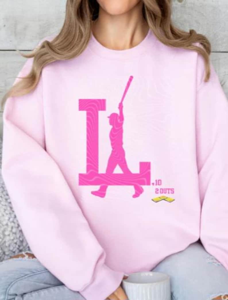 Op de afbeelding: Roze sweatshirt met een roze afbeelding van een honkballer die een bal slaat met de letter "L" op de achtergrond. De afbeelding bevat ook de tekst "10 OUTS".