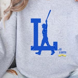 Op de afbeelding: Grijs sweatshirt met een blauwe afbeelding van een honkballer die een bal slaat met de letter "L" op de achtergrond. De afbeelding bevat ook de tekst "10 2 OUTS".