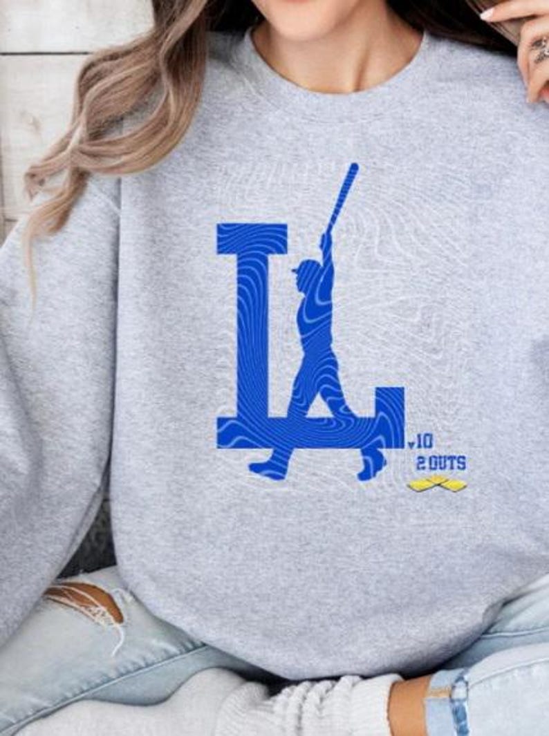 Op de afbeelding: Grijs sweatshirt met een blauwe afbeelding van een honkballer die een bal slaat met de letter "L" op de achtergrond. De afbeelding bevat ook de tekst "10 2 OUTS".