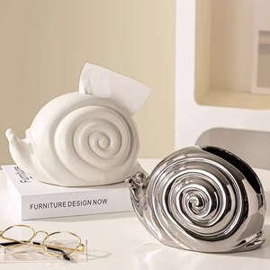 Puede incluir: Dos porta pa&ntilde;uelos de cer&aacute;mica con forma de caracol. Uno es blanco y el otro es plateado. El caracol blanco est&aacute; sobre un libro que dice "Furniture Design Now".