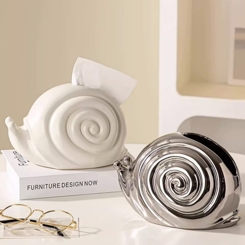 Puede incluir: Dos porta pa&ntilde;uelos de cer&aacute;mica con forma de caracol. Uno es blanco y el otro es plateado. El caracol blanco est&aacute; sobre un libro que dice "Furniture Design Now".