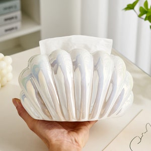 May include: White ceramic tissue box holder shaped like a seashell with iridescent detailing. The holder is open and has a white tissue inside.