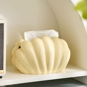 May include: A ceramic tissue box holder shaped like a seashell. The holder is a pale yellow colour and has a textured surface.