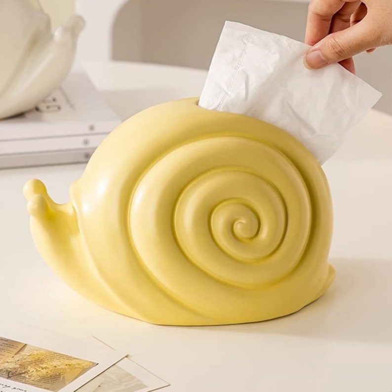 Puede incluir: Un porta pa&ntilde;uelos de cer&aacute;mica amarillo con forma de caracol. El porta pa&ntilde;uelos tiene una abertura redonda en la parte superior para los pa&ntilde;uelos.