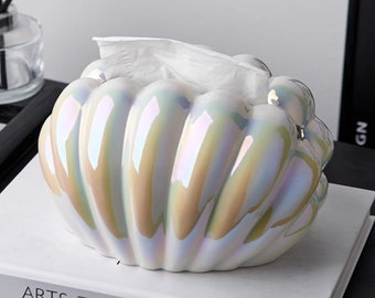 May include: A white ceramic tissue box holder shaped like a seashell with iridescent detailing. The holder is open and has a white tissue inside.