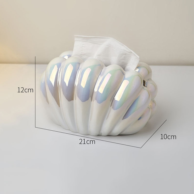 May include: White ceramic tissue box holder shaped like a seashell with iridescent detailing. The holder measures 21 cm long, 12 cm tall, and 10 cm wide.