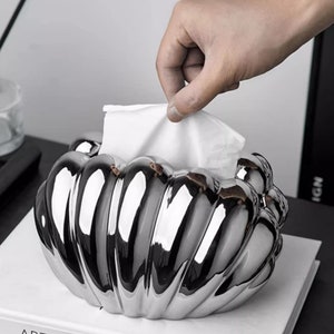 May include: A silver, ceramic tissue box holder shaped like a seashell. The holder has a glossy finish and is designed to hold a standard box of tissues.