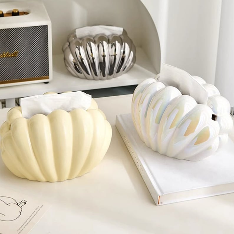 May include: Three ceramic tissue box covers in the shape of a pumpkin. One is silver, one is white with iridescent accents, and one is yellow.