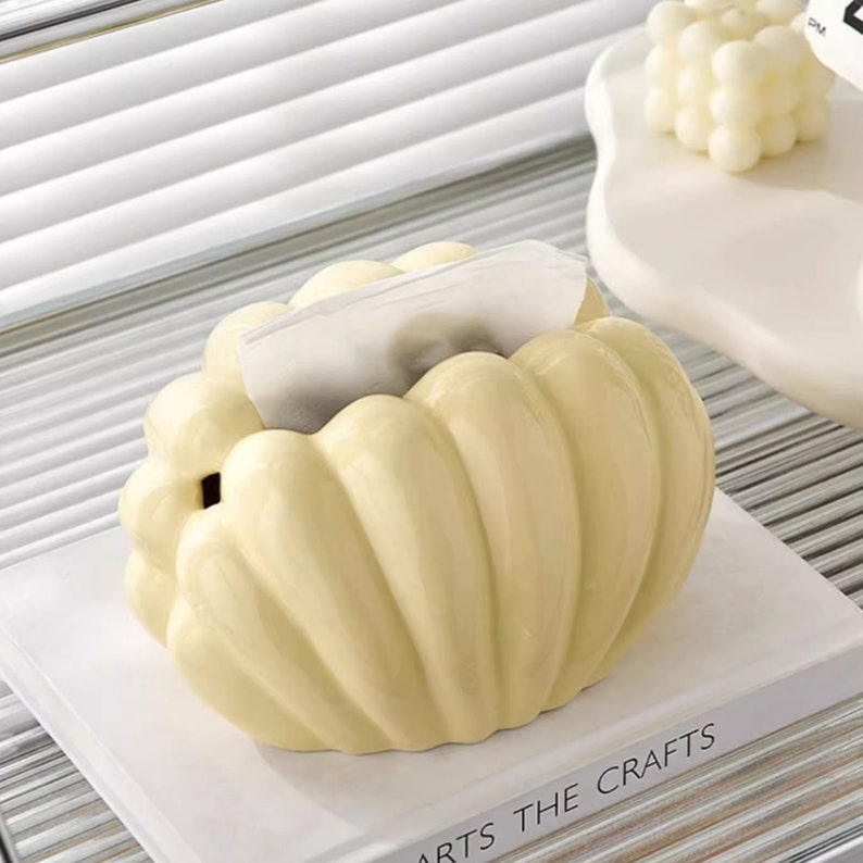 May include: A ceramic tissue box holder shaped like a seashell with a light yellow glaze. The holder has a textured surface and is designed to hold a standard tissue box.