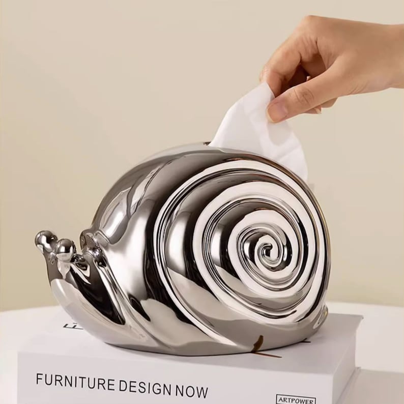 Puede incluir: Un porta pa&ntilde;uelos de tejido en forma de caracol plateado. El porta pa&ntilde;uelos est&aacute; hecho de un material met&aacute;lico brillante y tiene un dise&ntilde;o de espiral en la concha. Se est&aacute; sacando un pa&ntilde;uelo blanco del porta pa&ntilde;uelos.