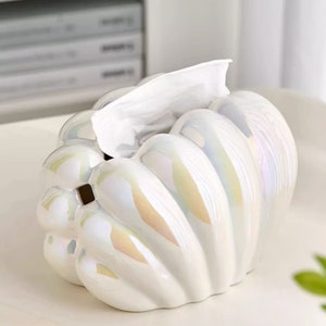 May include: White ceramic tissue box cover shaped like a seashell with iridescent detailing. The tissue box cover is empty and ready to be filled with tissues.