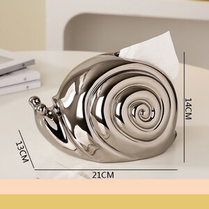 Puede incluir: Porta pa&ntilde;uelos de tejido en forma de caracol plateado con un pa&ntilde;uelo blanco en el interior. El porta pa&ntilde;uelos mide 21 cm de largo, 14 cm de alto y 13 cm de ancho.
