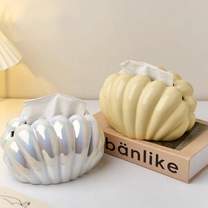 May include: Two ceramic tissue box covers shaped like seashells. One is white with iridescent detailing and the other is a pale yellow. Both are sitting on a table with a book that says "b&auml;nlike" on the cover.