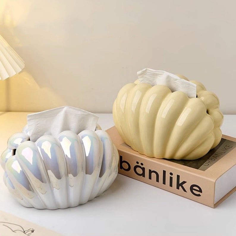 May include: Two ceramic tissue box covers shaped like seashells. One is white with iridescent detailing and the other is a pale yellow. Both are sitting on a table with a book that says "b&auml;nlike" on the cover.
