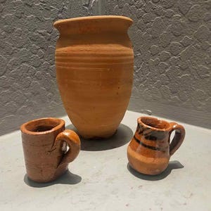 May include: A collection of rustic, handmade terracotta pottery. The set includes a large, unglazed vase and two small mugs with handles. The mugs have a brown and black striped design. The pottery has a warm, earthy tone.