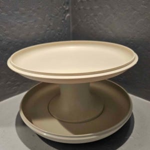 Puede incluir: Un soporte para servir de dos niveles, de color crema. El nivel superior es un plato circular elevado con un borde ligeramente elevado. El nivel inferior es un cuenco poco profundo más grande. Los dos niveles están conectados por un pedestal central cilíndrico.