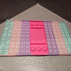 May include: A rectangular sensory toy in pastel colors. The toy features rows of circular bubbles in shades of mint green, pink, lavender, and a bright pink center. The bubbles are arranged in a grid pattern, designed for tactile interaction.