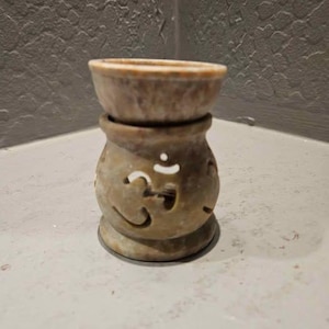 May include: A beige stone oil diffuser with a round base and a bowl-shaped top. The body features cut-out shapes and a carved Om symbol. The diffuser is designed to hold essential oils and is suitable for aromatherapy.