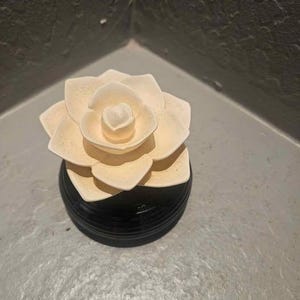 May include: A cream-colored, flower-shaped diffuser sits atop a black, round base. The flower has multiple layers of petals, creating a realistic appearance. The base has a glossy finish, and the overall design is elegant.