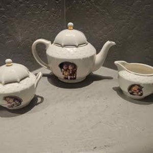May include: A white porcelain tea set featuring a teapot, sugar bowl, and creamer. Each piece has a textured surface and a small, colorful image of children. The teapot has a lid with a gold finial.